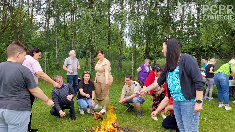 Zdjęcie: Grupa ludzi kuca i stoi przy ognisku i piecze kiełbaski na patykach. Są zadowoleni. Z tyłu widać stół i drzewa.
